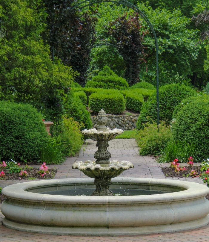 services-01 Beautiful stone fountain in a tranquil garden with lush green shrubs and colorful flowers in summer.